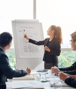 Attractive Asian businesswoman with ponytail pointing at diagram on marker board while holding working meeting in spacious boardroom