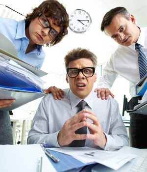 Pleading accountant looking at camera being surrounded by his partners with huge piles of documents